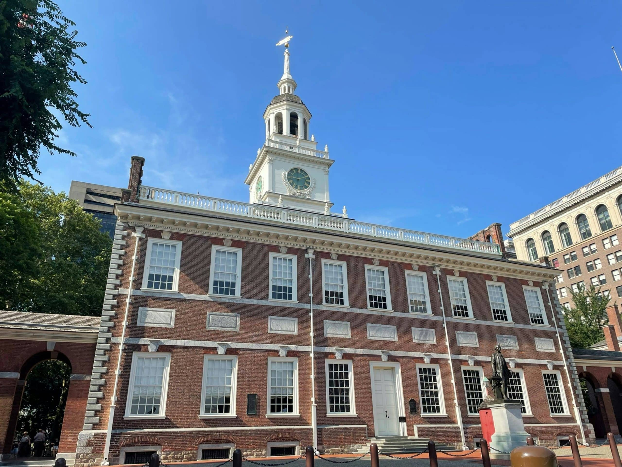 A historic red-brick building with white trim, large windows, and a tall clock tower stands under a clear blue sky—an iconic landmark that's expected to feature in Student Travel Trends for 2027, with a statue and trees gracing the foreground.