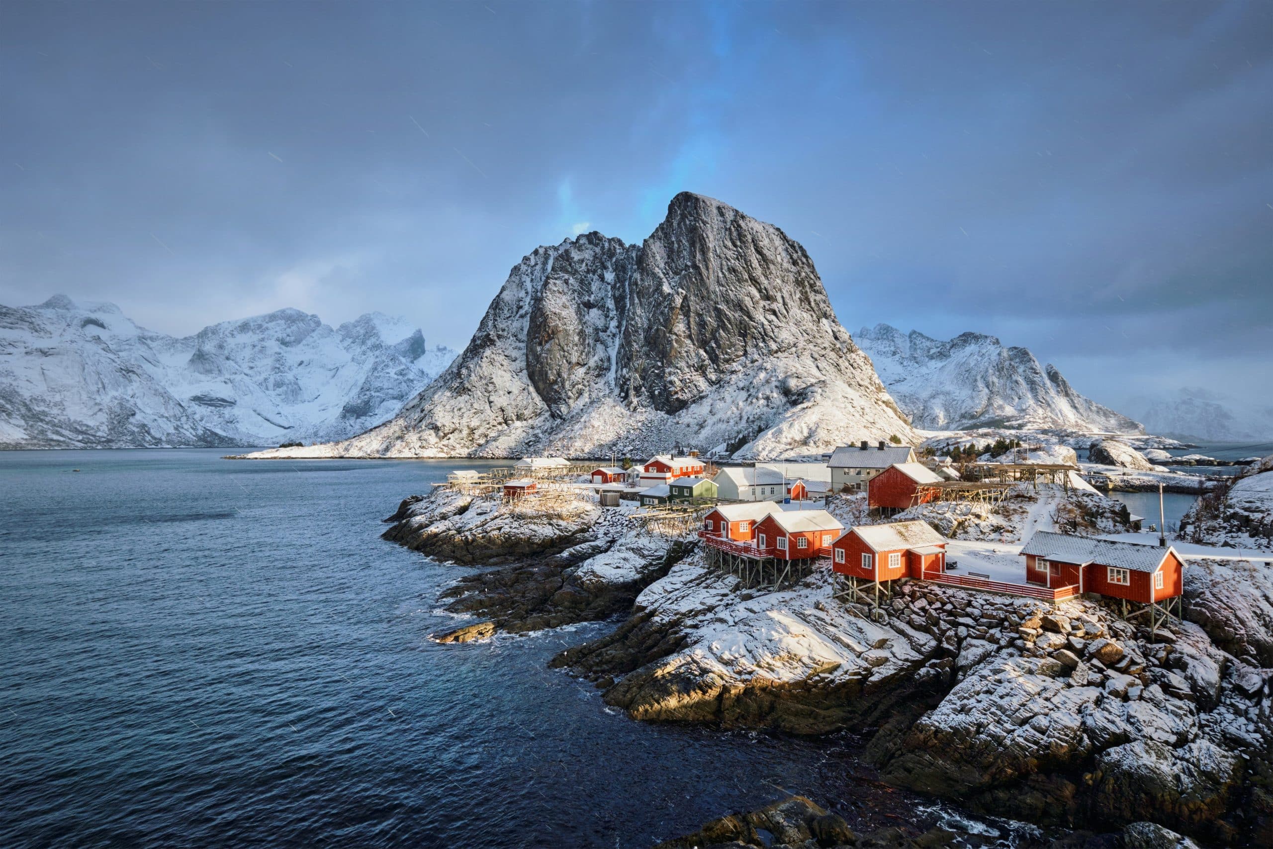 A coastal village with red cabins sits on a snowy, rocky shore—one of The Most Popular Student Travel Destinations for 2027—surrounded by calm blue water and dramatic, snow-covered mountains under a cloudy sky.