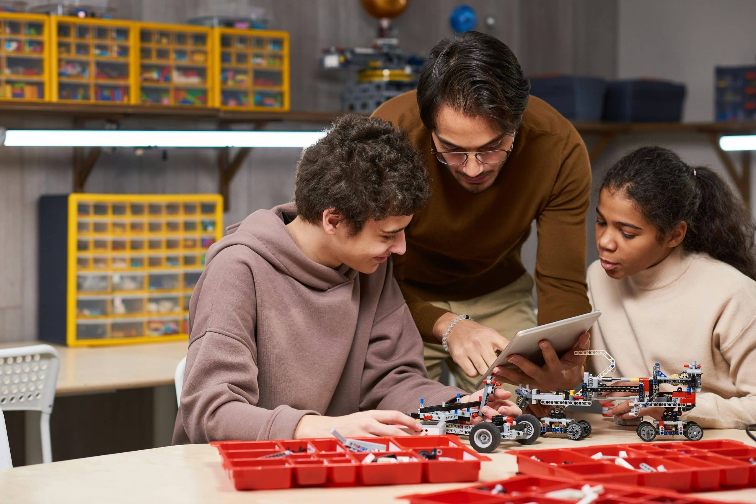 A teacher shows two students something on a tablet as they build robotic vehicles with LEGO parts in a classroom, making it a great STEM Washington DC school trip idea, surrounded by trays of building pieces and organized storage boxes.