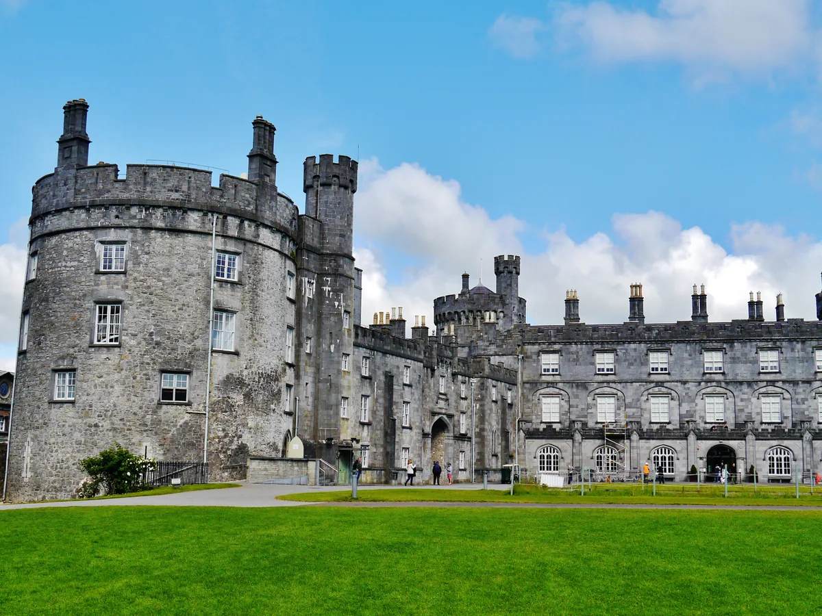 An Irish castle with a round tower and long stone range, seen across a lawn under a blue sky
