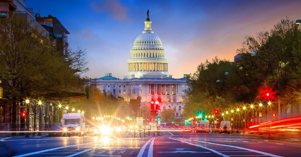 A nighttime view of a busy city street in Washington DC. The street lights are lit and the Capitol building is lit up.