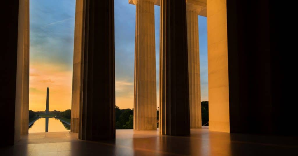 A sunset view of the Washington Monument and reflecting pond from the Lincoln Memorial steps. The pillars are sunlit.