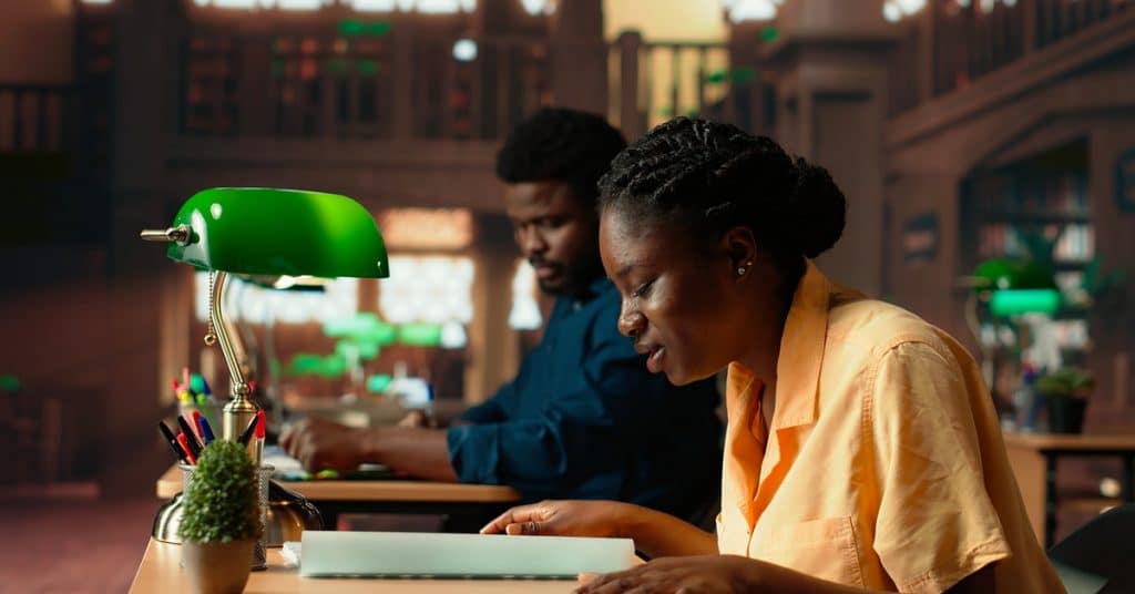 Two Black students sit in the library as they quietly study and read books on the table. There are green lamps.