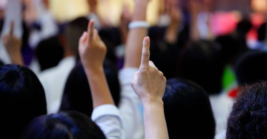 A crowd of people raises their hands, each showing one finger pointed upward, suggesting participation, voting, or agreement in a group setting.