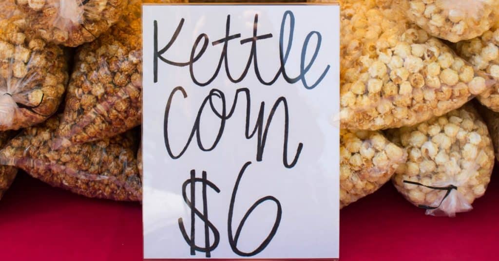 A handwritten sign reads "Kettle Corn $6" in front of several bags of kettle corn displayed on a red tablecloth.