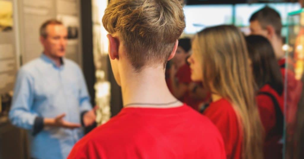 A group of high school students stand close to an exhibit on a field trip as a professional discusses the details.