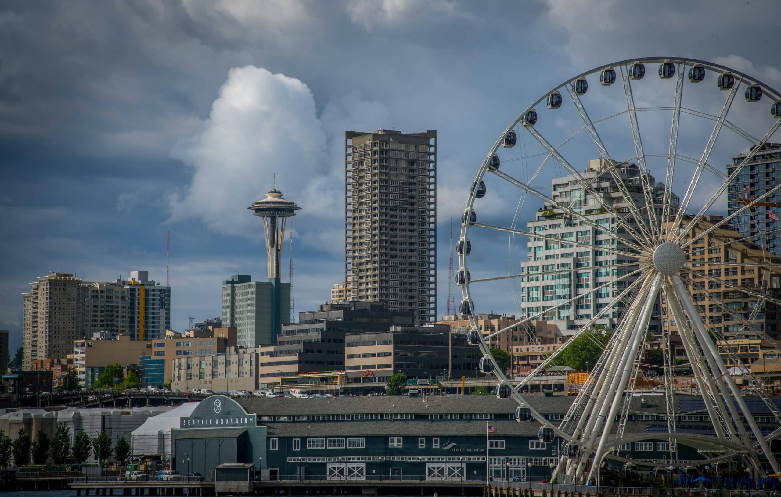 Seattle waterfront with the Seattle Great Wheel in the foreground, the Space Needle and tall buildings in the background, and dramatic clouds in the sky—a must-see among the top 10 things to do on a Seattle field trip.