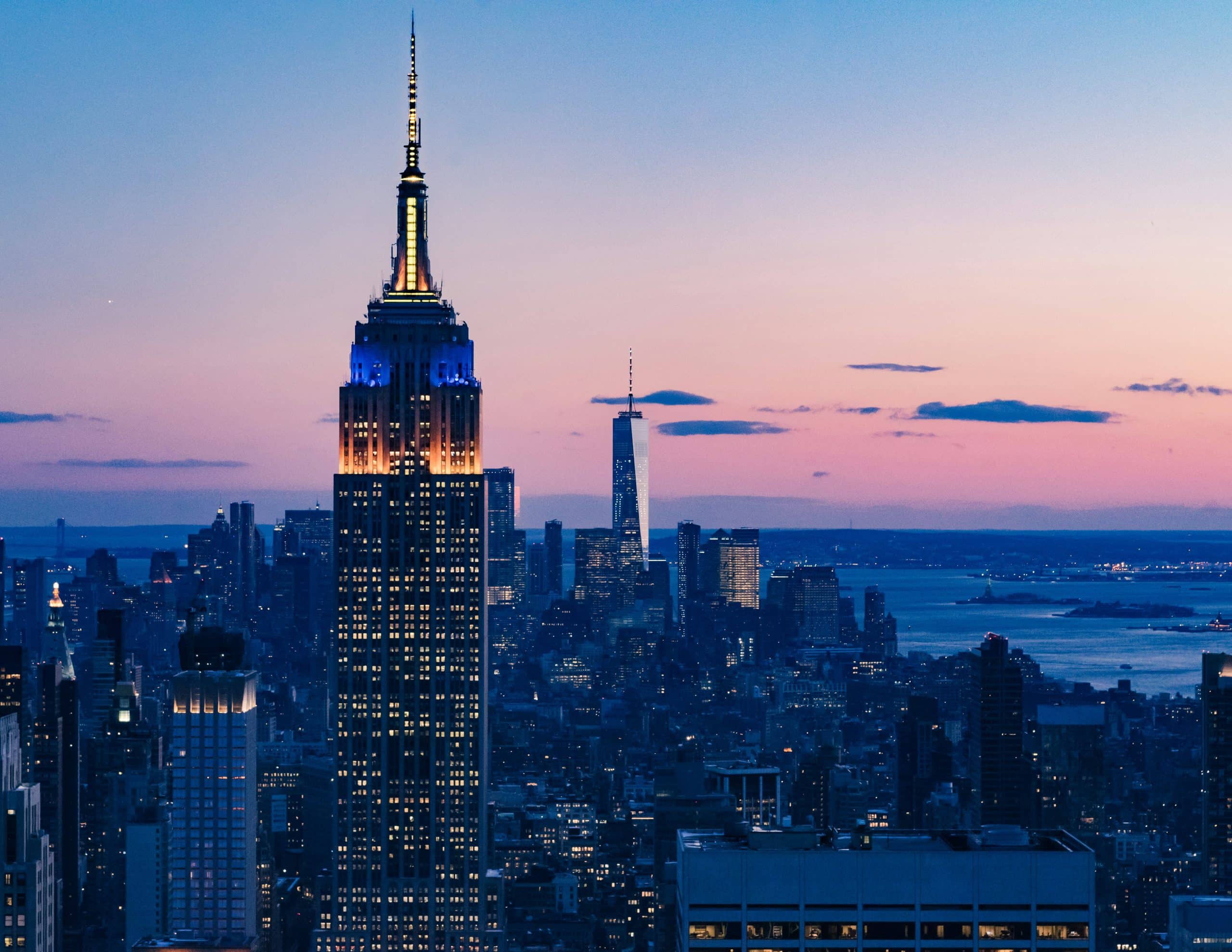 The Empire State Building, one of The Most Popular Student Travel Destinations for 2027, is illuminated at dusk with blue and gold lights, standing tall among the New York City skyline as pink and purple hues color the sky.