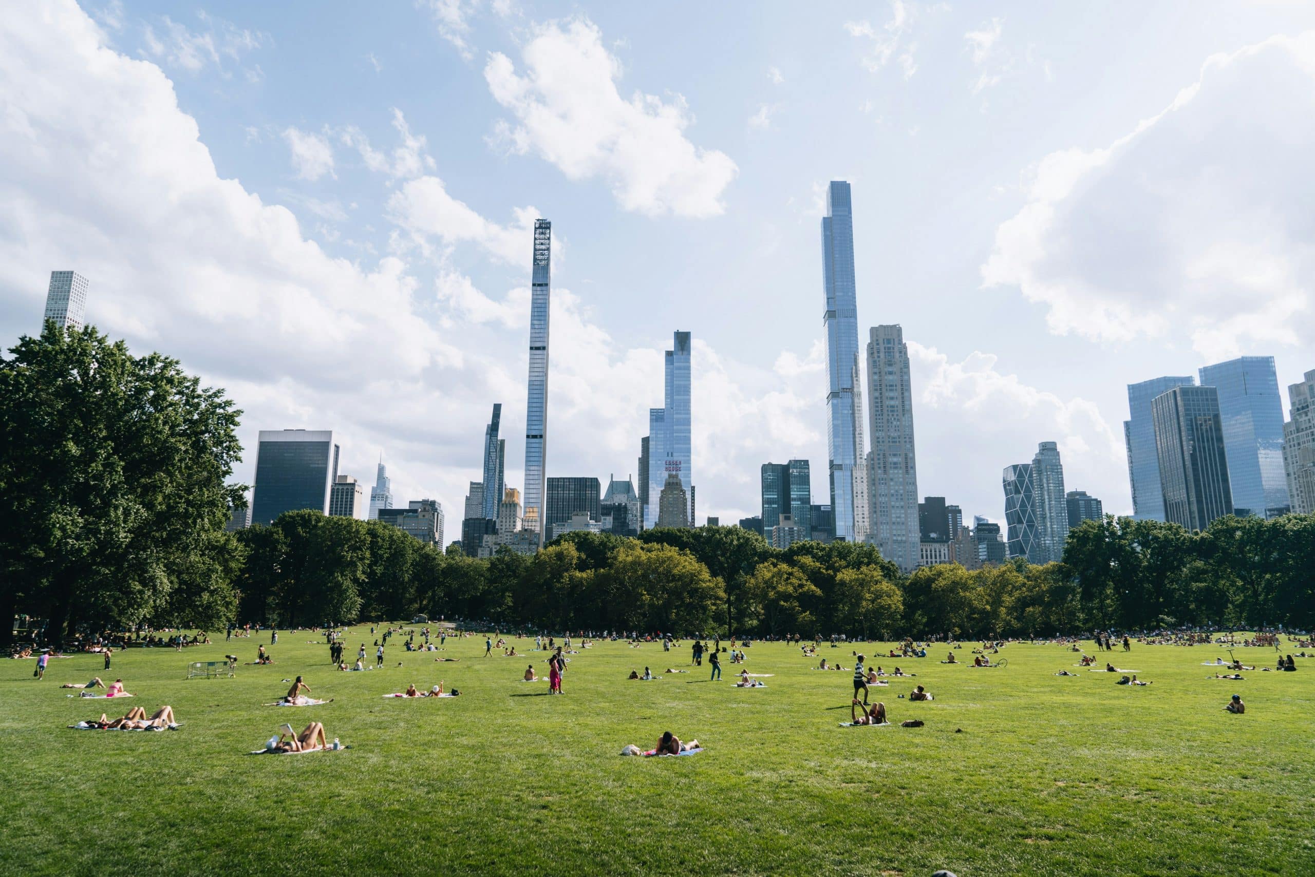 People relax and sunbathe on a large grassy field in a city park, with tall skyscrapers and trees in the background under a partly cloudy sky.