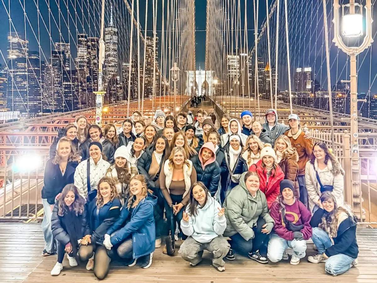 A large Landmark student group photographed on the Brooklyn Bridge at night with the Manhattan skyline lit behind them
