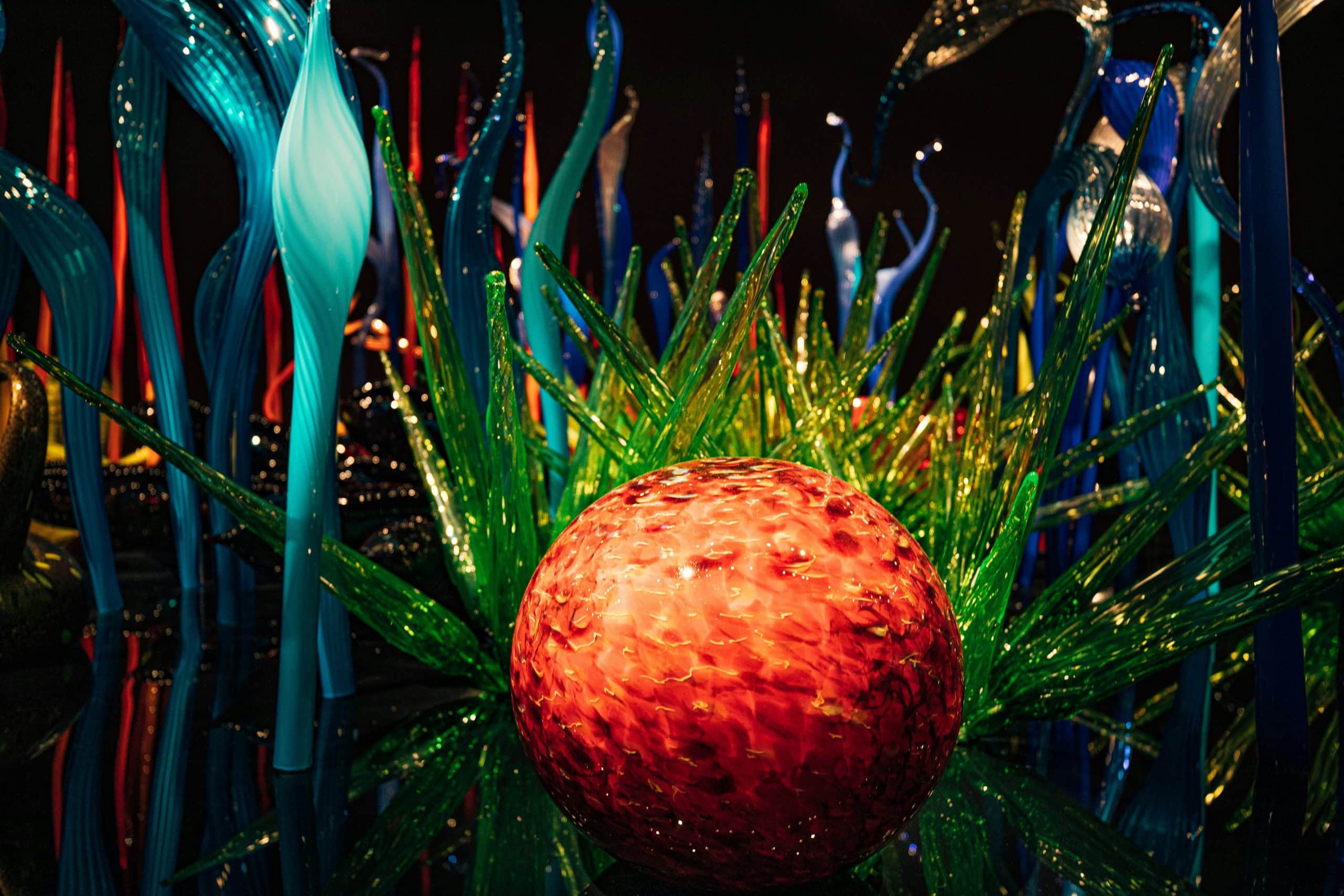 A vibrant glass art display from a Seattle School Trip, featuring a large, textured red orb surrounded by spiky green glass shapes and tall, twisted glass sculptures in blue, orange, and clear hues on a dark background.