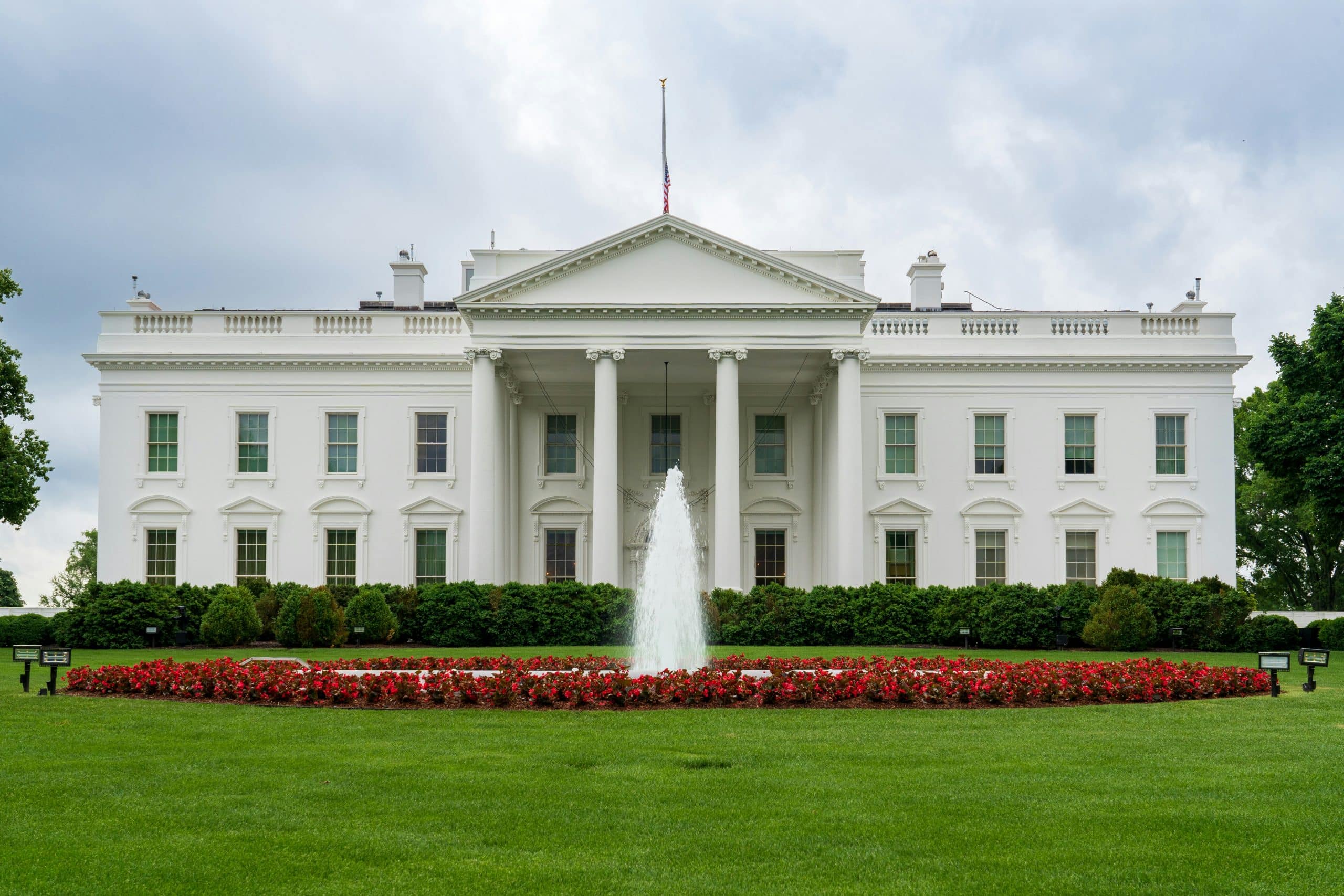 The White House, a large white neoclassical building with columns, stands behind a fountain and red flowers on a green lawn under a cloudy sky—an iconic destination for educational field trips to Washington DC.