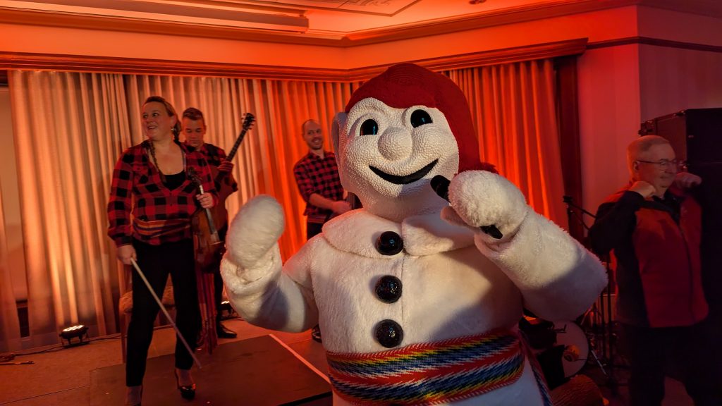 A person in a large white mascot costume with a red hat and multicolored belt poses cheerfully. Behind them, musicians in red and black attire play instruments on a small stage, under warm lighting.