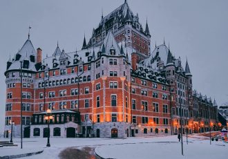 Fairytale like castle with turrets and intricate architecture, surrounded by snow. The building is illuminated warmly against a gray sky, creating a contrast with the snowy foreground. A few people are seen walking nearby.