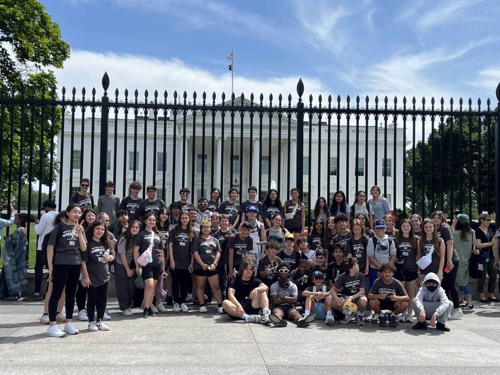 A large group of people, mostly wearing matching shirts, pose in front of the White House behind a black iron fence. The sky is clear and sunny, and the American flag is visible atop the building.