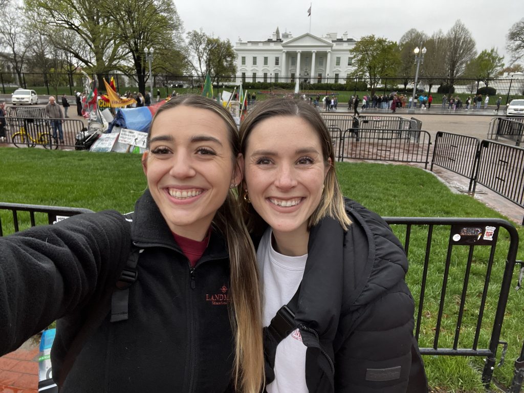 Two people taking a selfie in front of the White House on a cloudy day. They are smiling and standing close together, with barricades and green grass behind them. The White House is visible in the background.