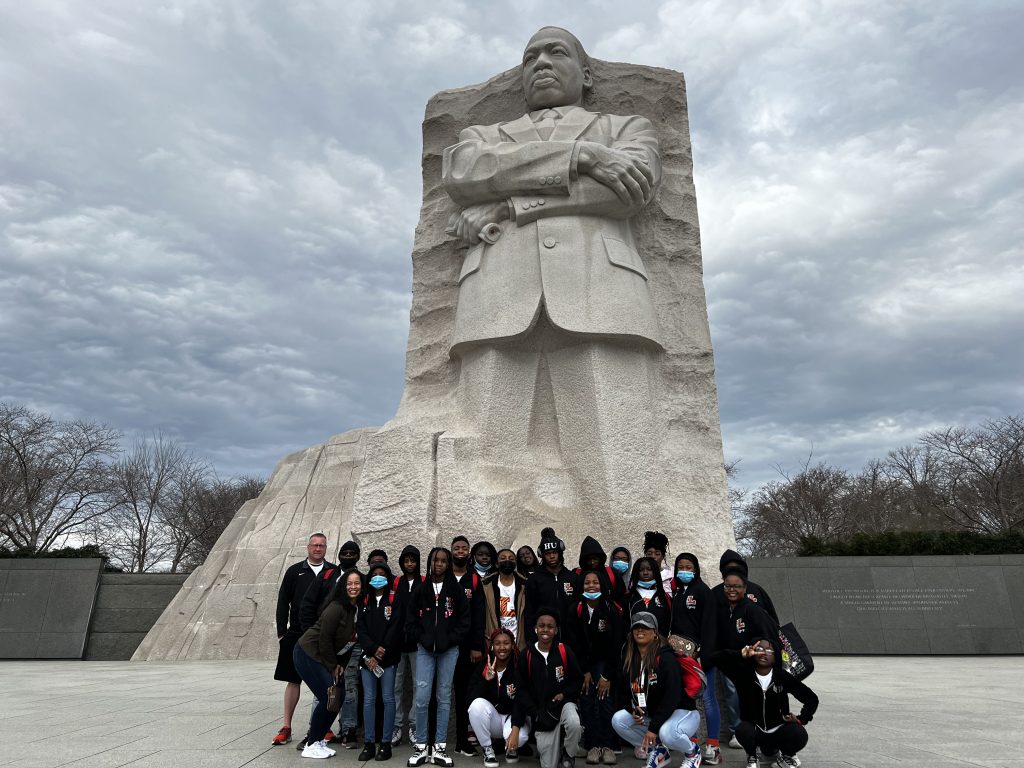 A group of people posing in front of the Martin Luther King Jr. Memorial in Washington, D.C. The monument features a large stone statue of Martin Luther King Jr. with his arms crossed, against a cloudy sky backdrop.