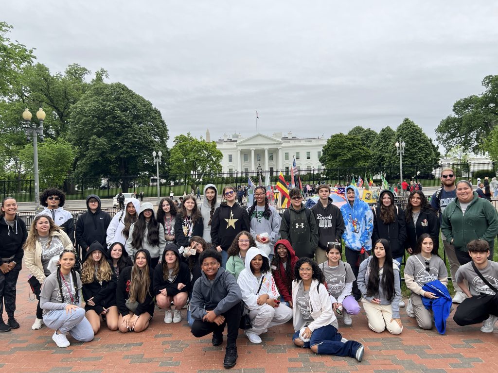 A large group of people pose for a photo in front of the White House. They are dressed in casual clothing, and some hold colorful flags. Trees and cloudy skies are in the background.