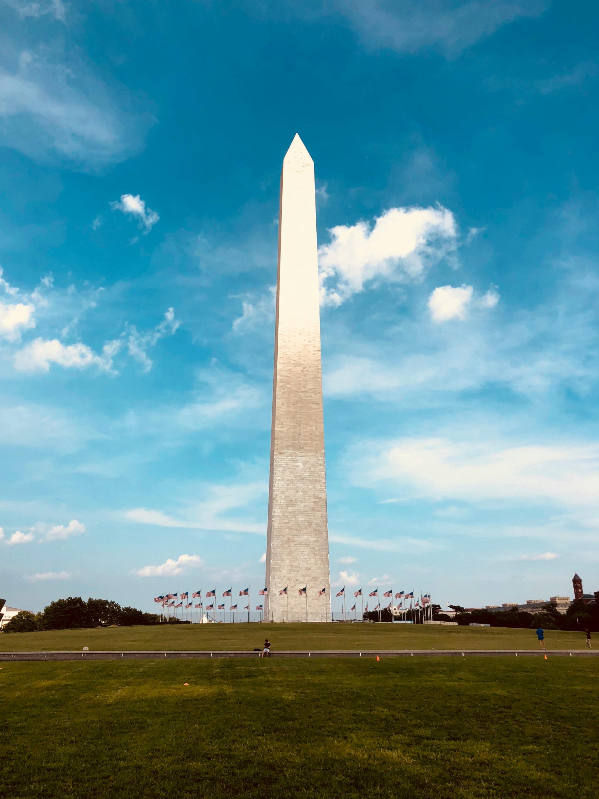 The image shows the Washington Monument, a tall, white obelisk, under a clear blue sky with scattered clouds. The structure is surrounded by a circle of flags and a grassy area.