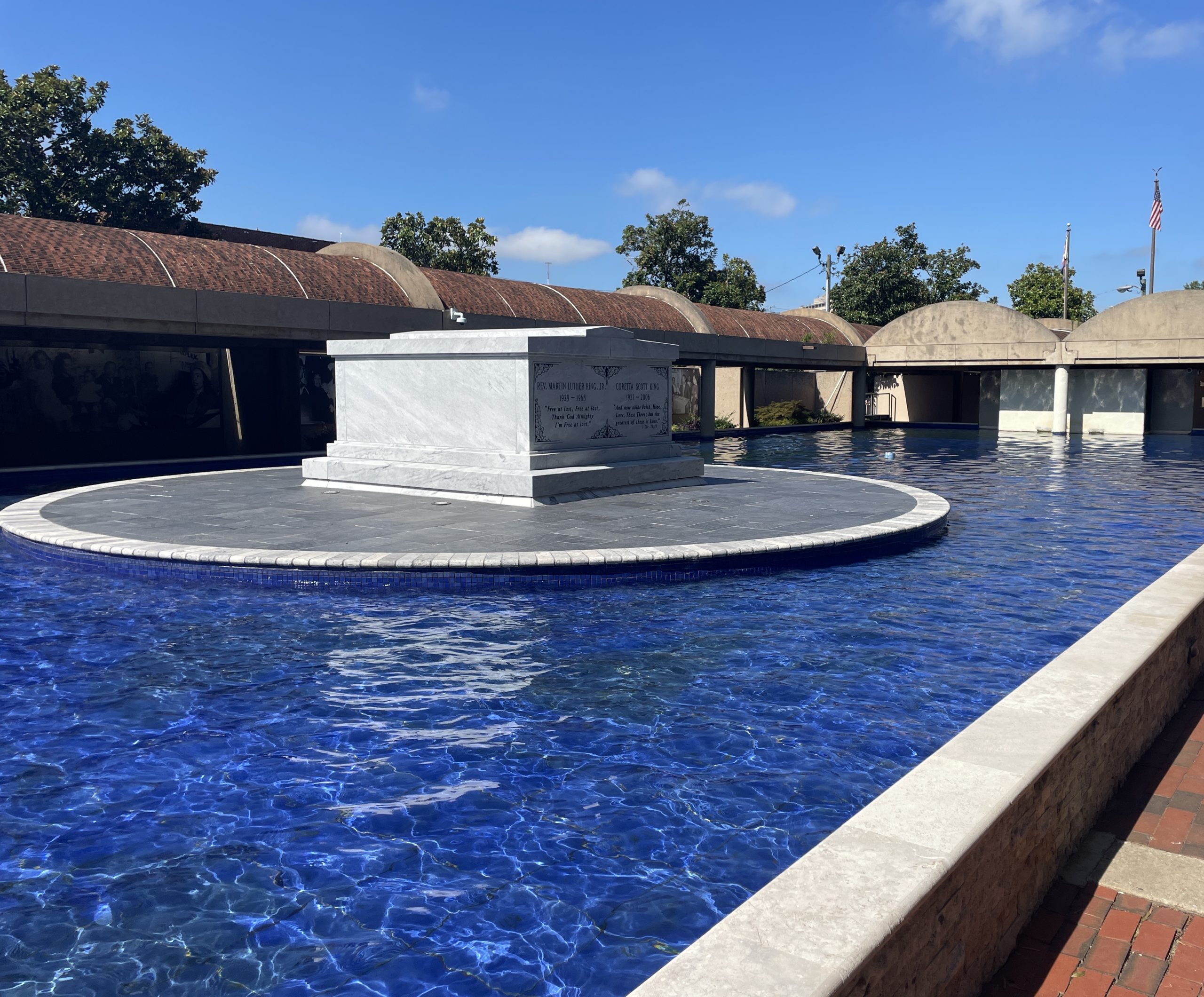 A white marble tomb sits on a circular platform at the heart of Martin Luther King Jr. National Historical Park, surrounded by a deep blue reflecting pool, with trees and a low, curved building beyond under a clear sky.