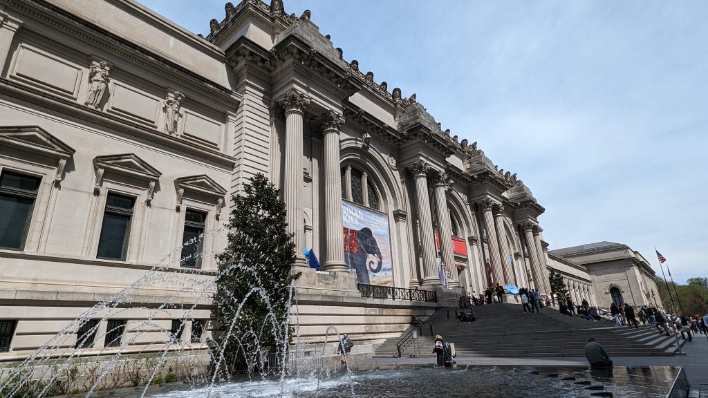 Wide-angle view of the Metropolitan Museum of Art’s grand neoclassical facade, a water fountain in the foreground, people on the steps, and banners by the entrance—capturing what to see at the Metropolitan Museum of Art under a partly cloudy sky.