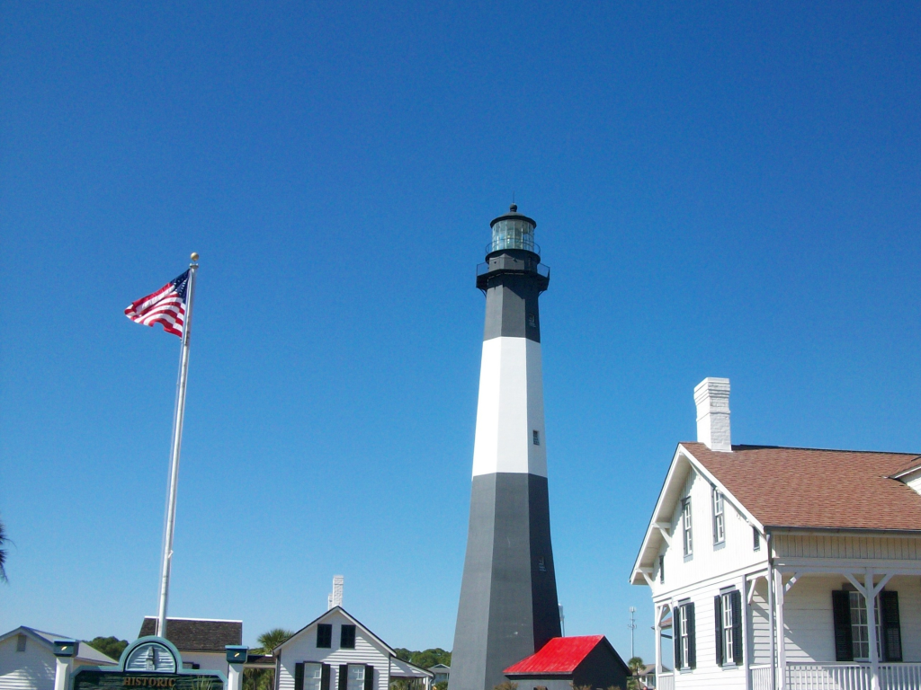 A tall black and white striped lighthouse stands under a clear blue sky, surrounded by white buildings and an American flag—a perfect site for Landmark Educational Tours registration.