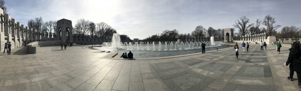 Wide-angle view of the World War 2 Memorial Washington DC, featuring a large fountain at the center, tall stone pillars, arches, and people walking or sitting around the plaza.