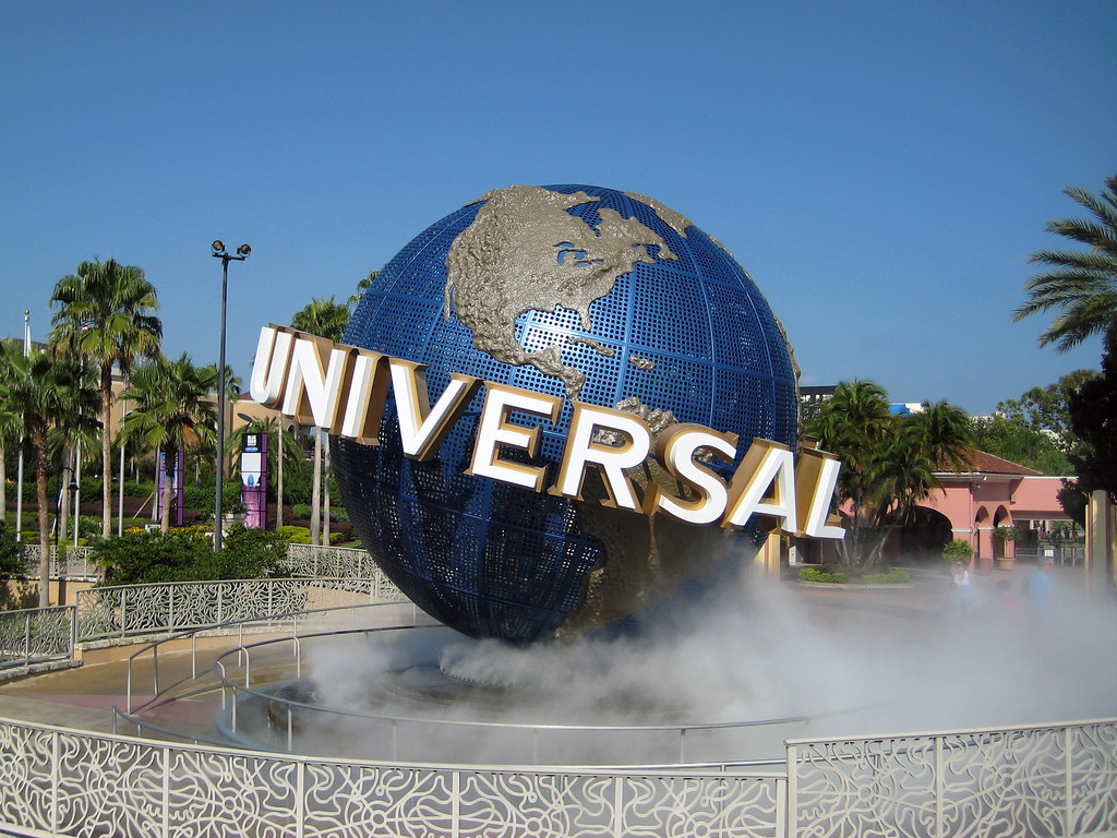 A large blue globe with the word "Universal" encircling it stands as a landmark at the entrance of Universal Studios, greeting visitors on their 3 Day Orlando Student Trip, surrounded by mist, palm trees, and a clear blue sky.