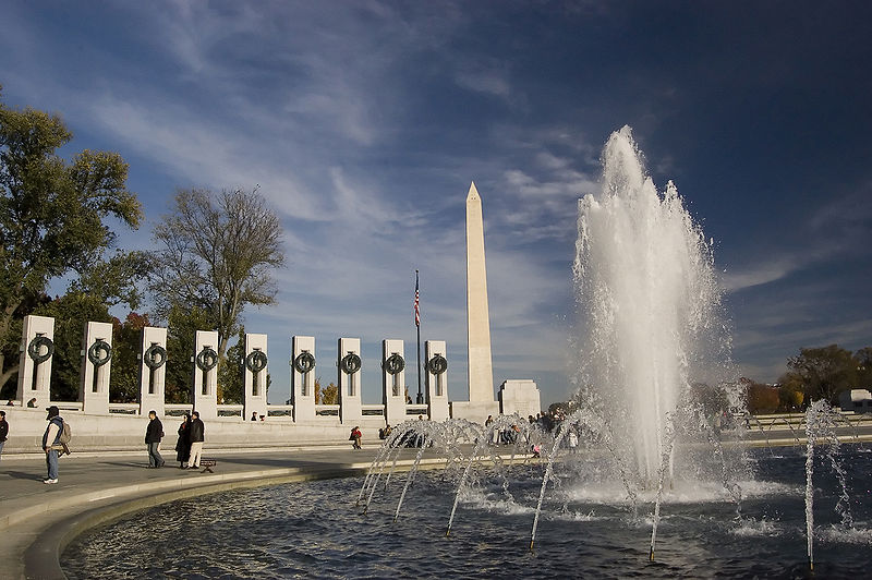 People walk around the World War II Memorial in Washington, D.C., with a large fountain in the foreground and the Washington Monument rising in the background—an ideal spot to visit during an Evening Monument Tour DC under a blue sky.