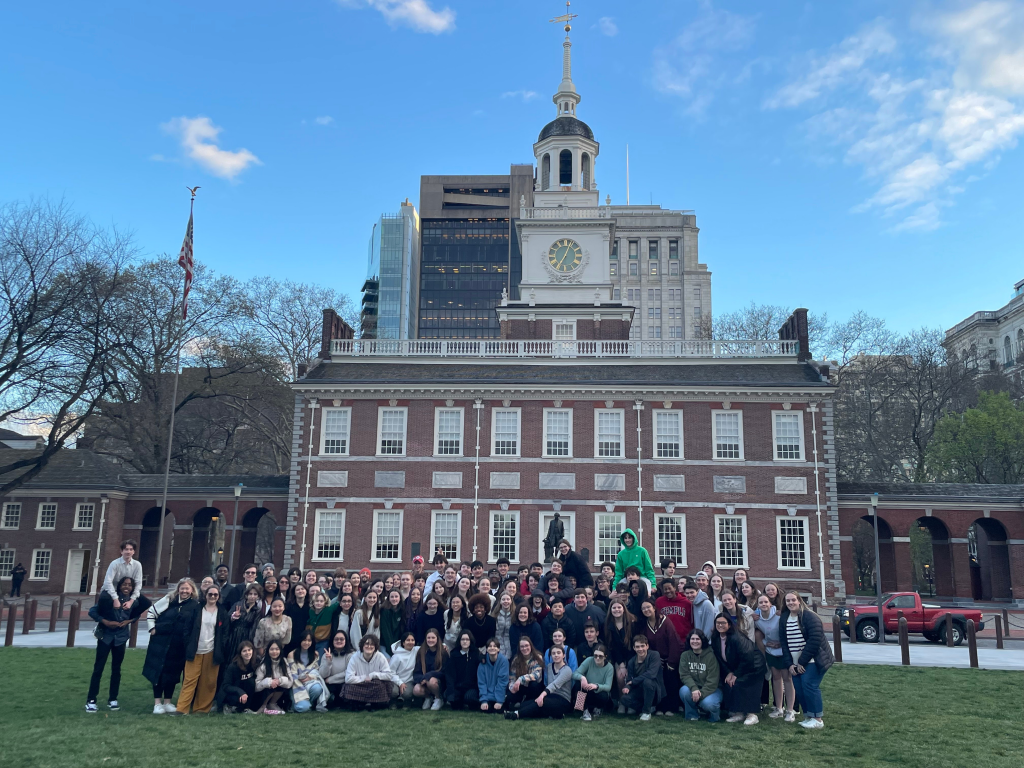 A large group of people poses in front of Independence Hall, a historic red-brick building with a clock tower, on a grassy lawn under a partly cloudy sky.