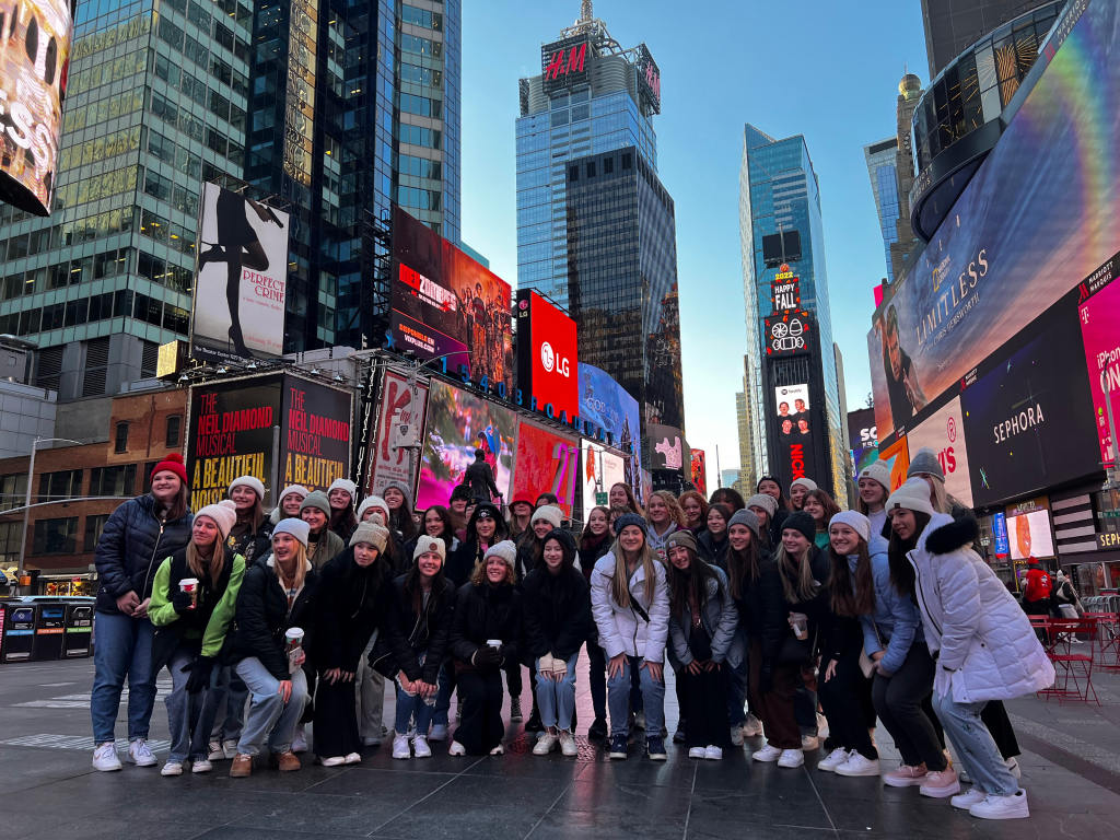 A large group of smiling people dressed in winter clothing pose together in Times Square, New York City, surrounded by tall buildings and colorful electronic billboards.