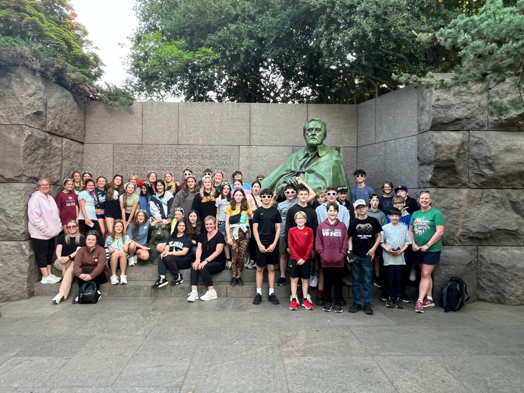 A large group of people, mostly teens and a few adults, pose in front of a statue of Franklin D. Roosevelt at an outdoor monument surrounded by stone walls and greenery.
