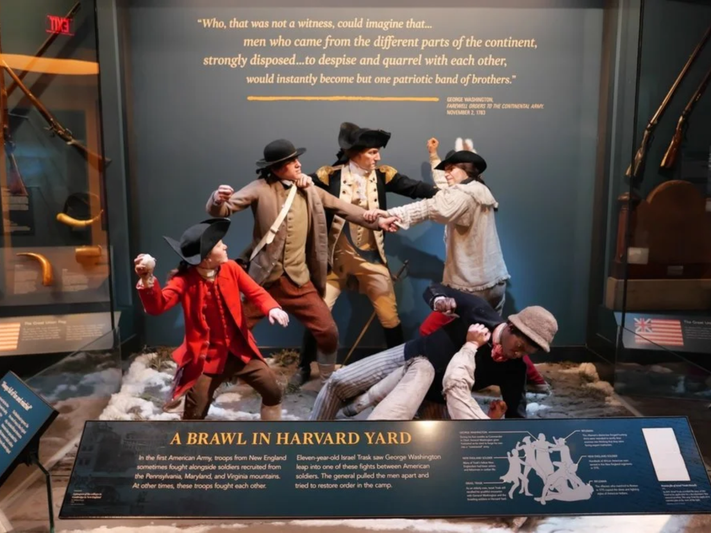 A museum exhibit shows five life-sized mannequins dressed as Revolutionary War soldiers fighting. Some are grabbing or punching each other. Historical rifles and a quote are displayed in the background.