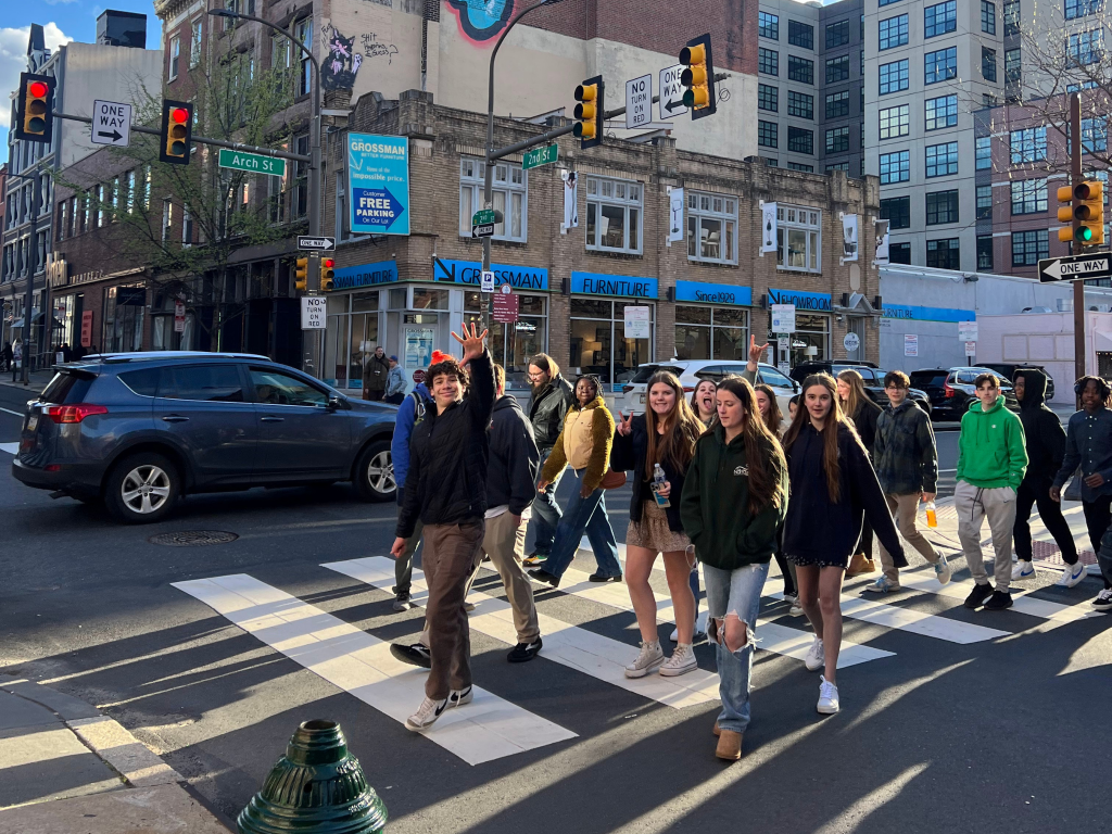 A group of smiling young people cross a busy city street at a crosswalk, surrounded by cars and tall buildings. One person waves at the camera, and the sun casts long shadows on the pavement.
