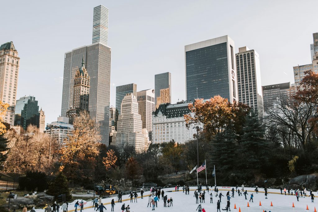 People ice skating at an outdoor rink in Central Park, New York City, with tall skyscrapers and autumn trees in the background under a clear sky. Book your winter NYC trip 2026 to experience this magical scene firsthand!.