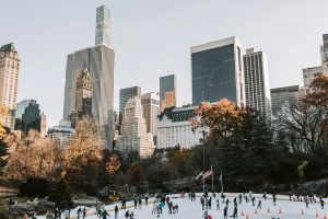 People ice skating at an outdoor rink in Central Park, New York City, with tall skyscrapers and autumn trees in the background under a clear sky. Book your winter NYC trip 2026 to experience this magical scene firsthand!.