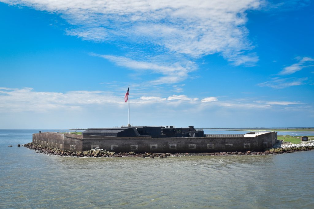 A historic brick fort sits on an island surrounded by water under a blue sky with scattered clouds. Perfect for Charleston School Field Trips, the site features an American flag atop the fort and green grass behind the structure.