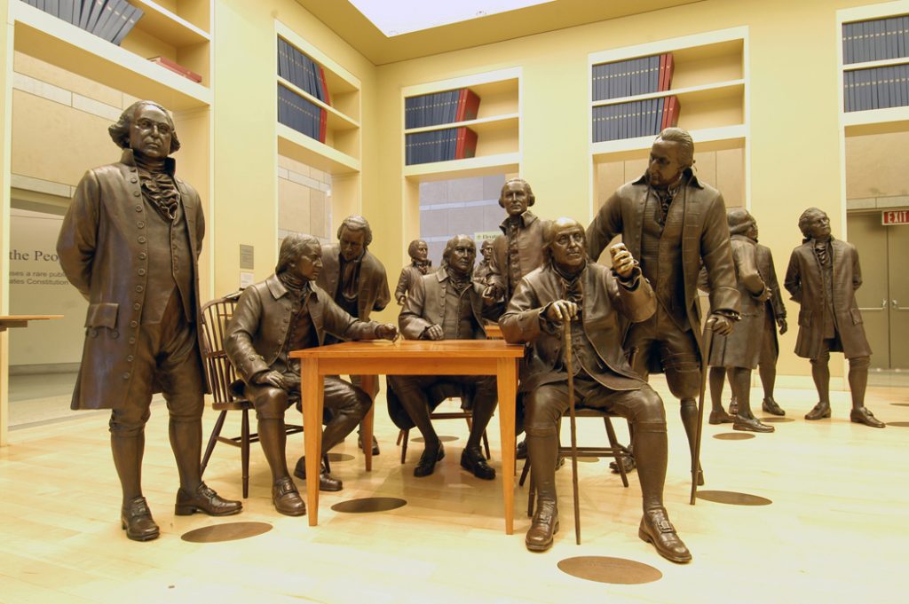 Lifelike bronze statues of several men in period clothing gather around a wooden table in a brightly lit room with bookshelves, recreating a scene as timeless as the iconic moments celebrated in Historic MLB Ballparks.