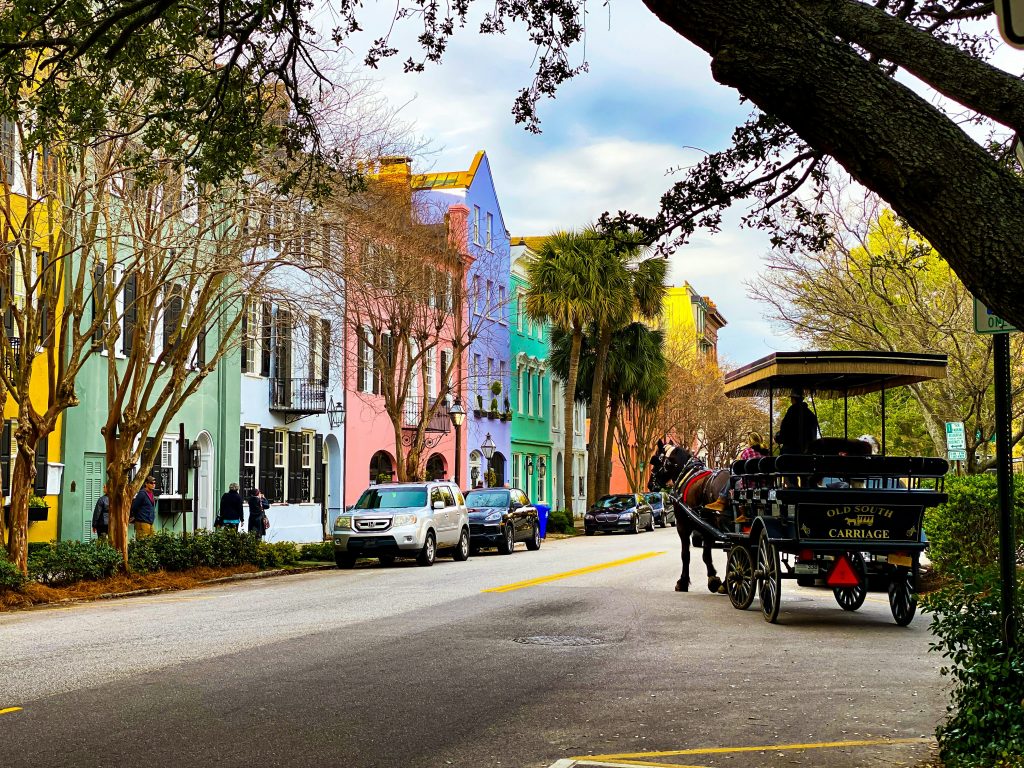A row of colorful pastel houses lines a street with parked cars, trees, and a horse-drawn carriage carrying passengers—a charming sight often enjoyed on Charleston School Field Trips under a partly cloudy sky.