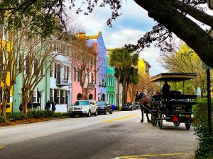 A row of colorful pastel houses lines a street with parked cars, trees, and a horse-drawn carriage carrying passengers—a charming sight often enjoyed on Charleston School Field Trips under a partly cloudy sky.