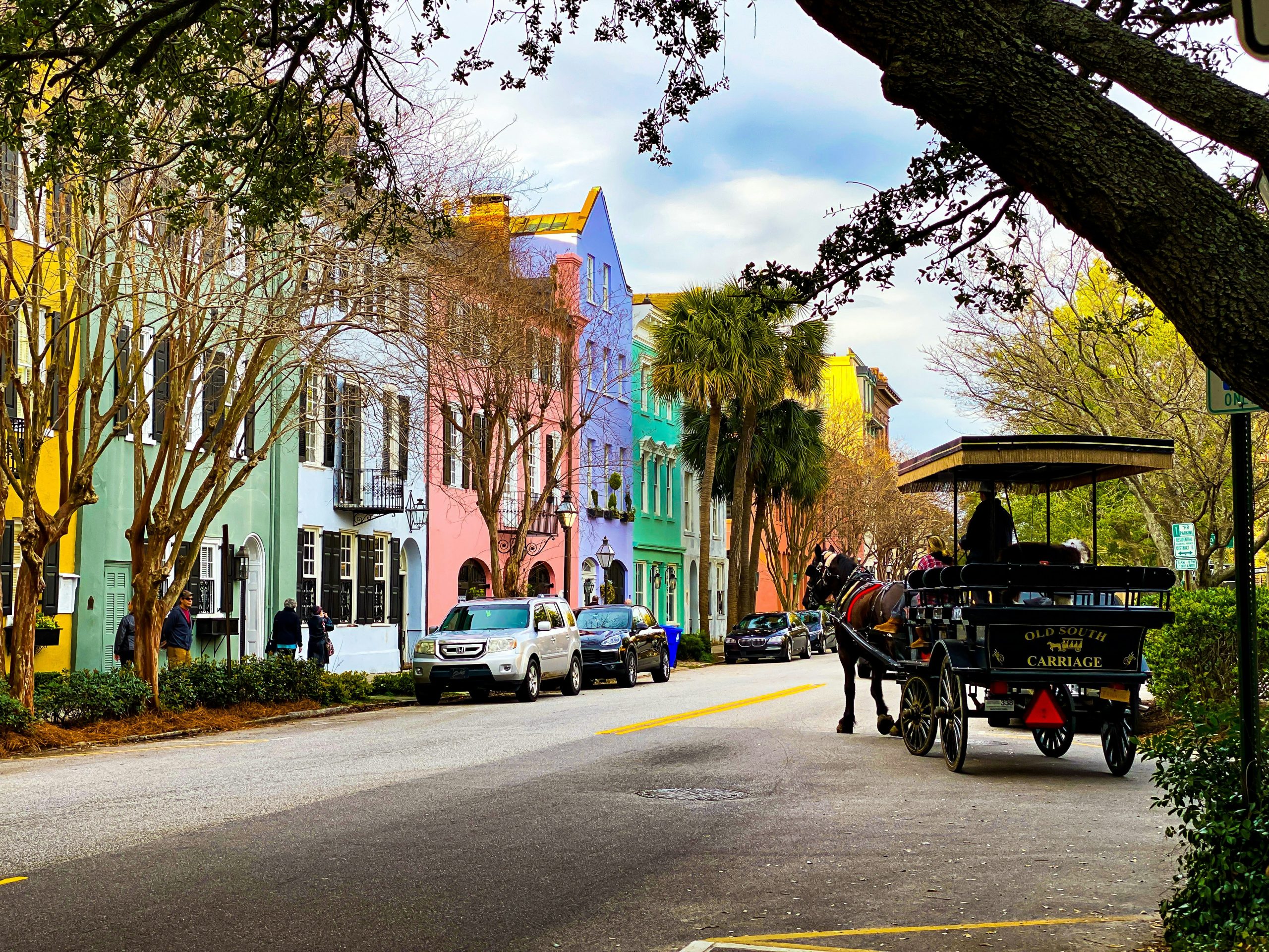 A row of colorful pastel houses lines a street with parked cars, trees, and a horse-drawn carriage carrying passengers—a charming sight often enjoyed on Charleston School Field Trips under a partly cloudy sky.