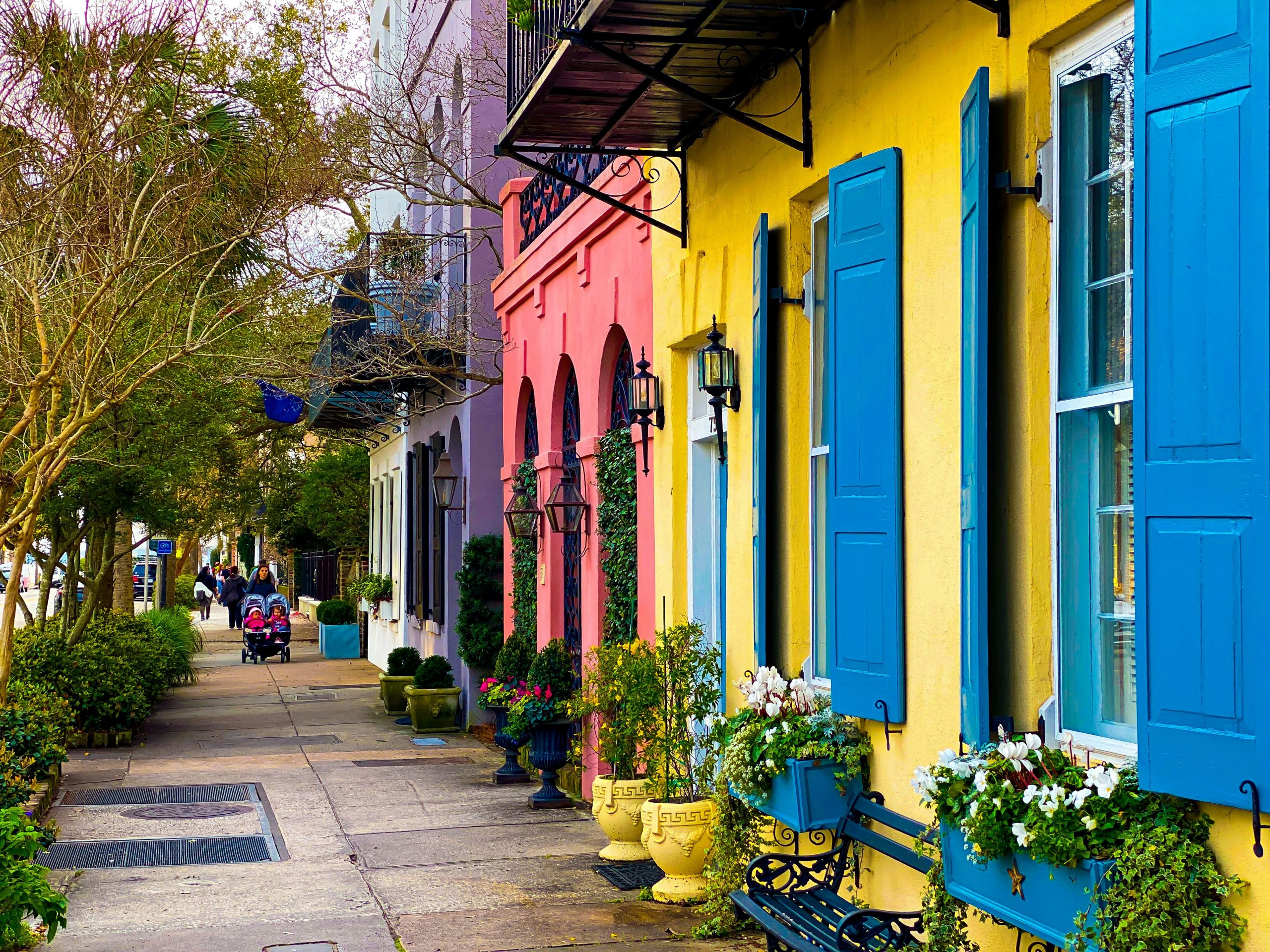 A colorful street scene with pastel buildings in yellow, pink, and purple, each with bright blue shutters and flower boxes—perfect for capturing the charm of a Charleston STEM class trip as students stroll down the plant-lined sidewalk.