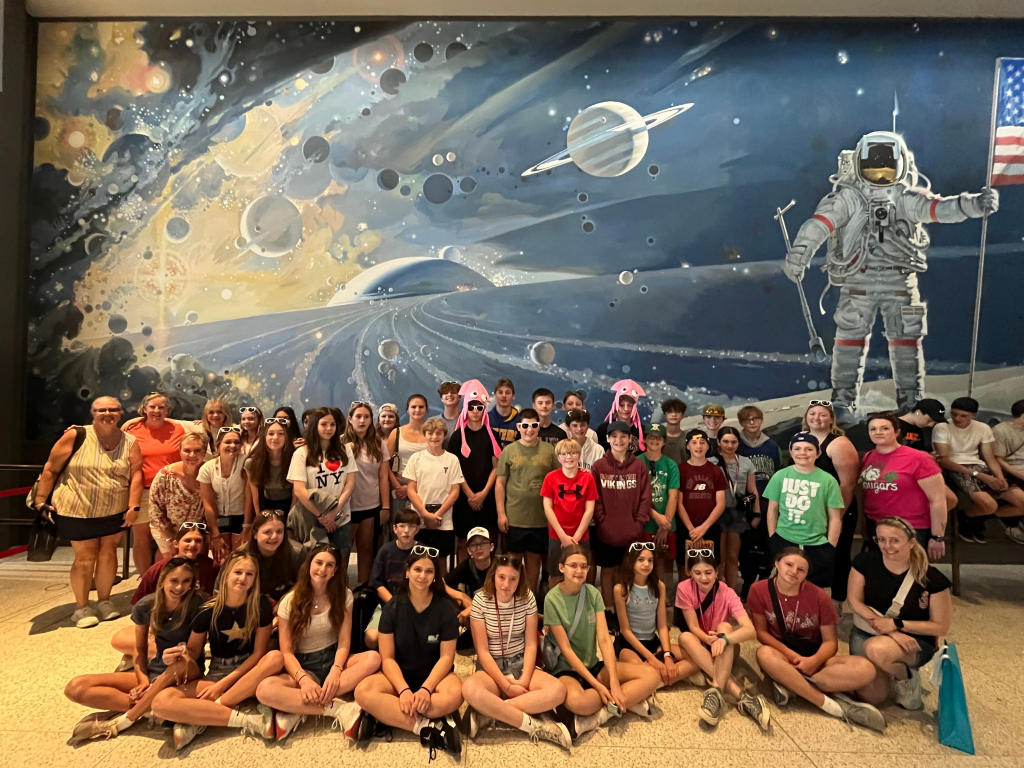 A large group of children and adults pose in front of a colorful space-themed mural featuring an astronaut holding an American flag and planets in the background. Some kids wear sunglasses and novelty glasses.