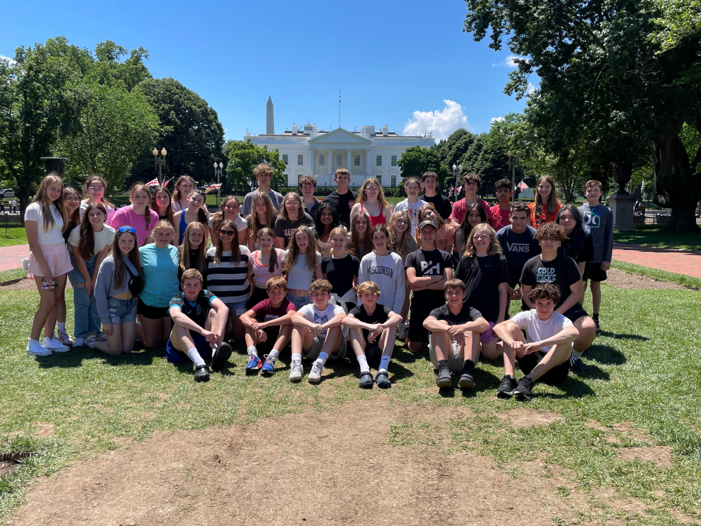 A large group of teens poses for a photo on a sunny day in front of the White House, with the Washington Monument visible in the background and trees surrounding them.