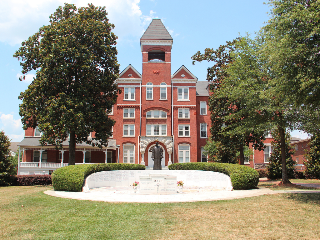 A historic red-brick building with arched windows and a central tower stands behind a curved white stone memorial engraved with names, surrounded by green lawns, trees, and shrubs under a blue sky.