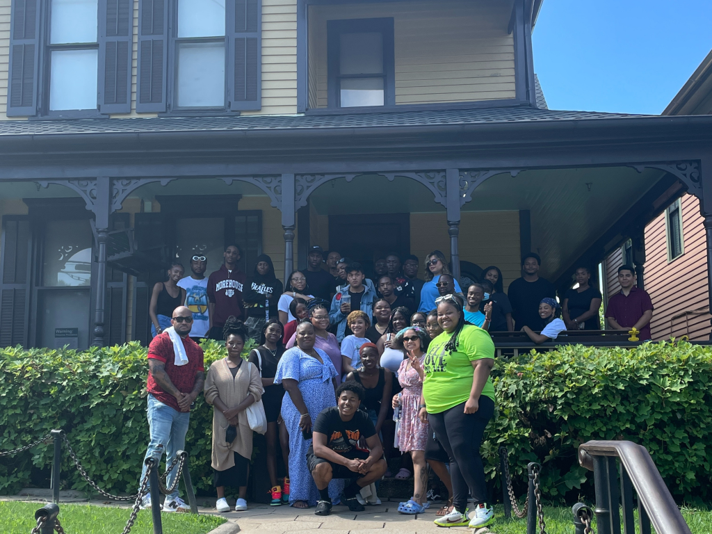 A large group of people pose together, smiling for a group photo in front of a historic yellow house with a porch on a sunny day. Some people stand on the porch while others are on the steps and walkway.