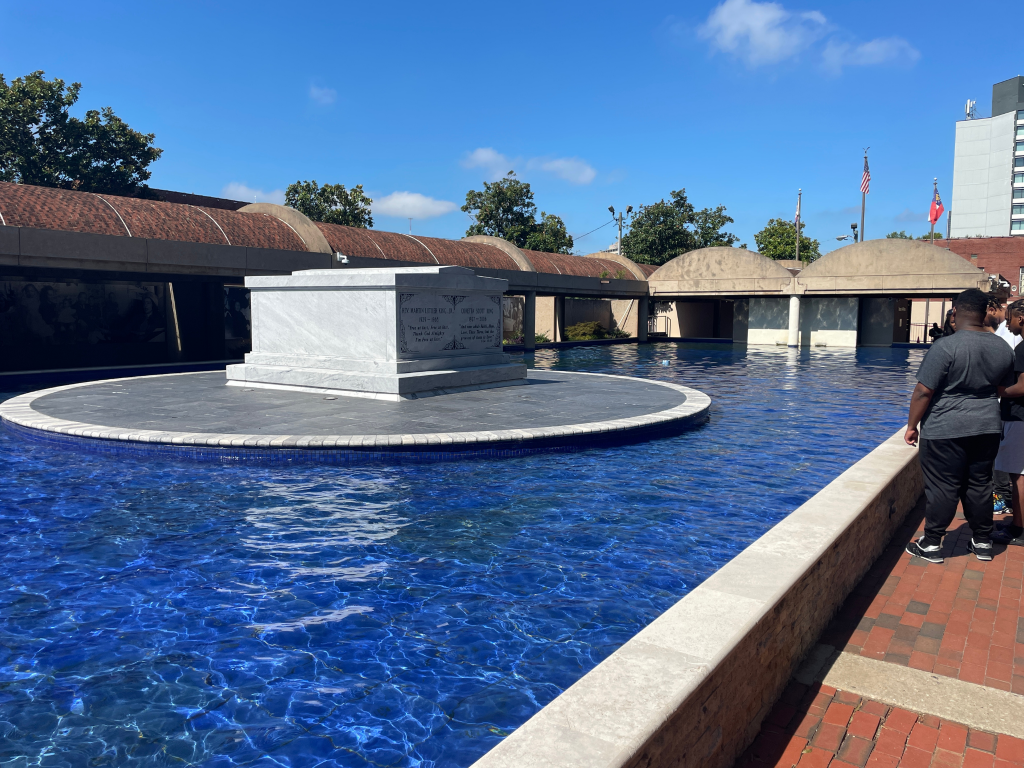 A white stone tomb sits on a circular platform surrounded by a blue reflecting pool. People stand nearby on a brick walkway, and trees and flagpoles are visible in the background under a clear sky.