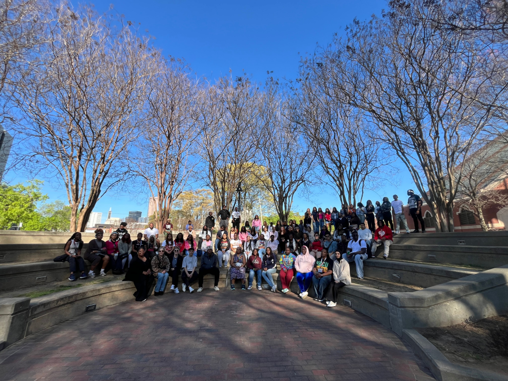 A large group of people sit and stand on tiered concrete steps in an outdoor amphitheater, surrounded by leafless trees under a clear blue sky.