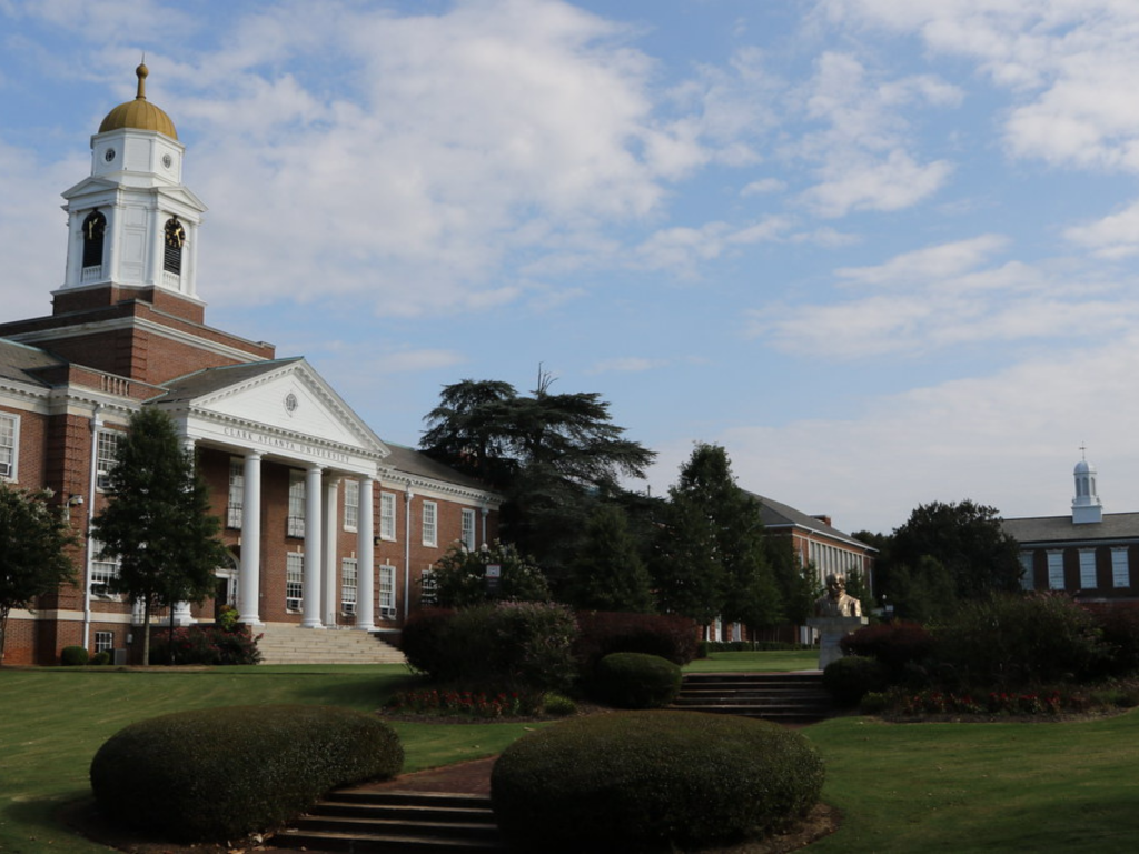 A large brick academic building with white columns and a clock tower, surrounded by green lawns, trees, and bushes under a partly cloudy sky.