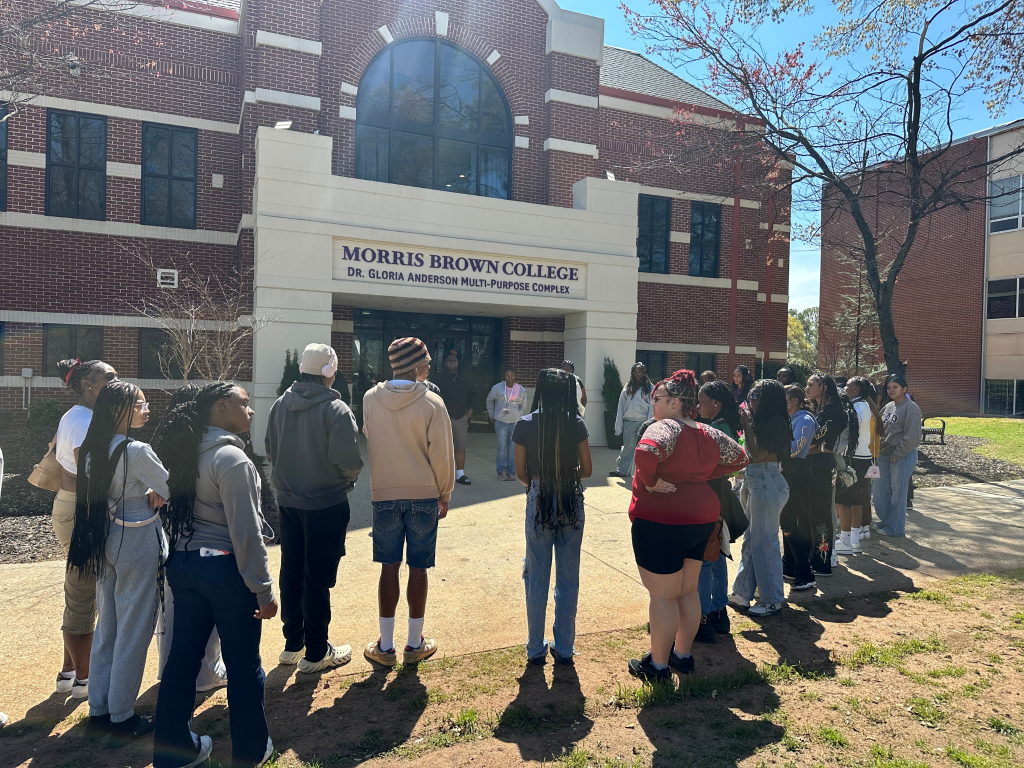 A group of students stands in front of the Dr. Gloria Anderson Multi-Purpose Complex at Morris Brown College, listening to a person speak during a sunny day.